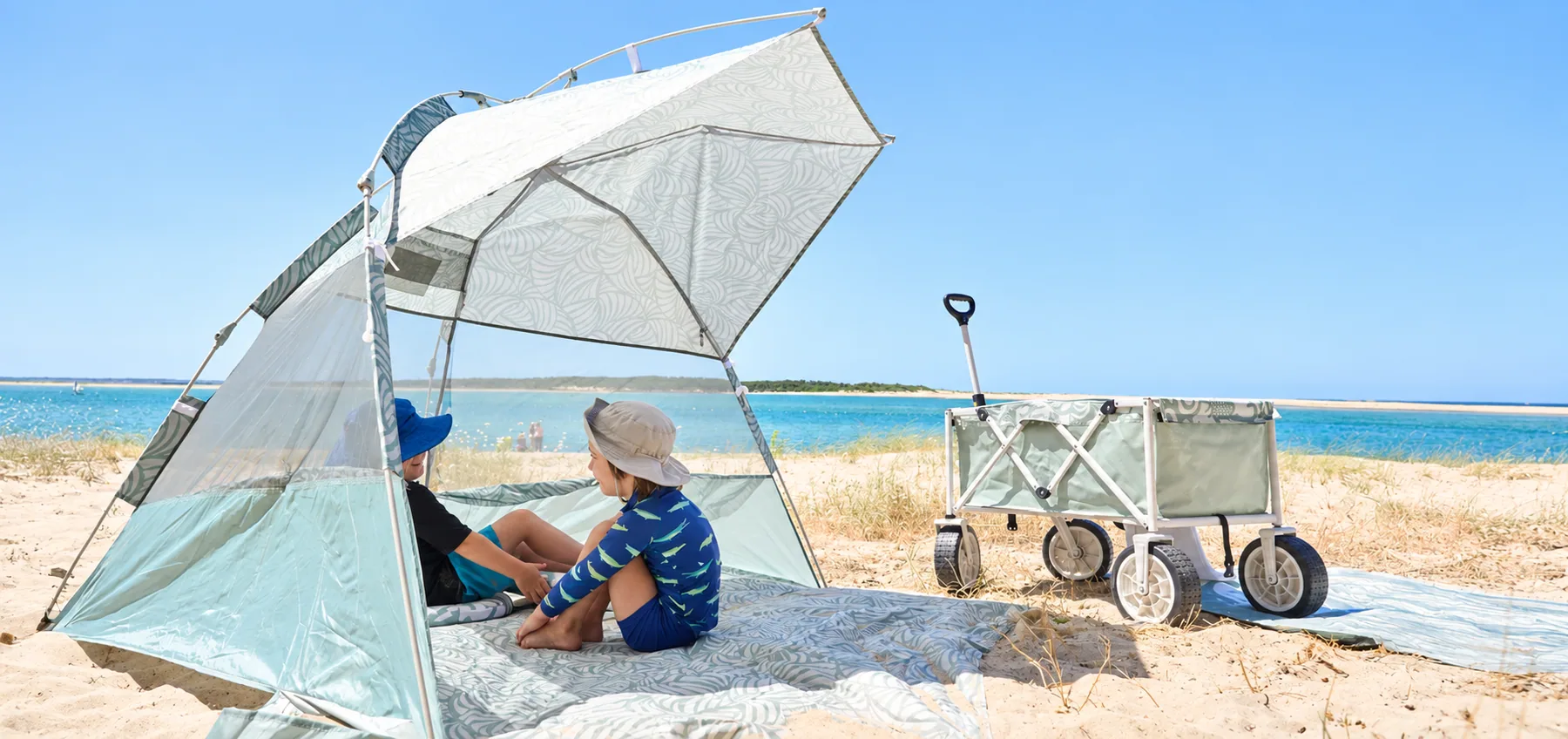 A sage beach wagon beside a patterned shelter with two children relaxing on the sand by the sea